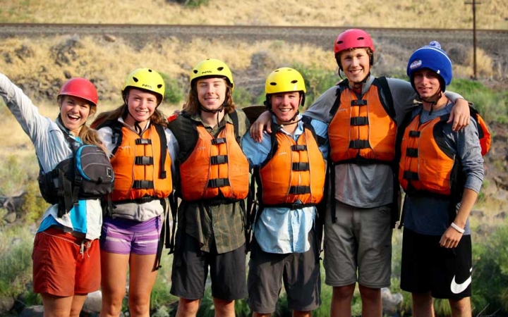 Six people wearing helmets and life jackets stand on land, wrap their arms around each other and smile at the camera.